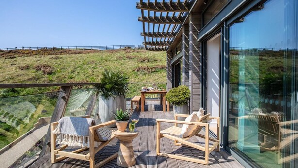 En træterrasse med udendørs møbler, et pergola og en glasvæg med udsigt over en bakke.