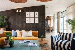 A modern living room with a wooden wall, a sofa, striped armchair, and a coffee table with books and plants. - 4 Bed in Watergate Bay (oc-b584453) (Tregurrian)