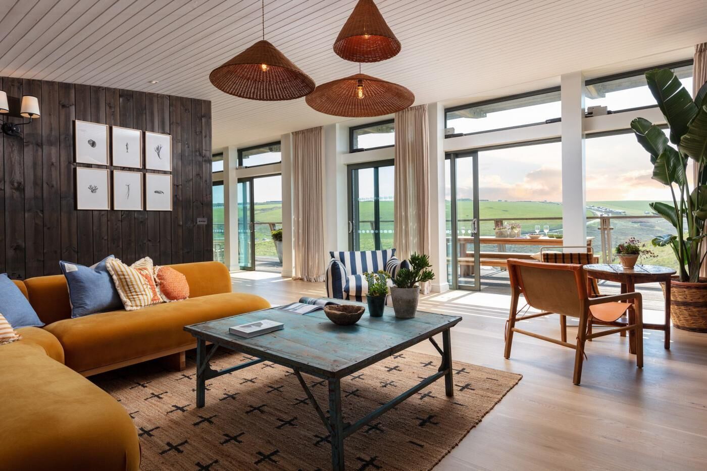 A modern living room with a sofa, coffee table, and dining area, featuring large windows and a view of the countryside.