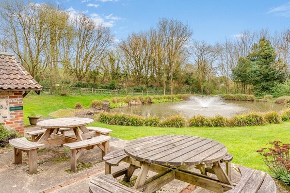 Un espace pique-nique avec des tables et des bancs en bois, une petite construction en briques et un étang agrémenté d’une fontaine.