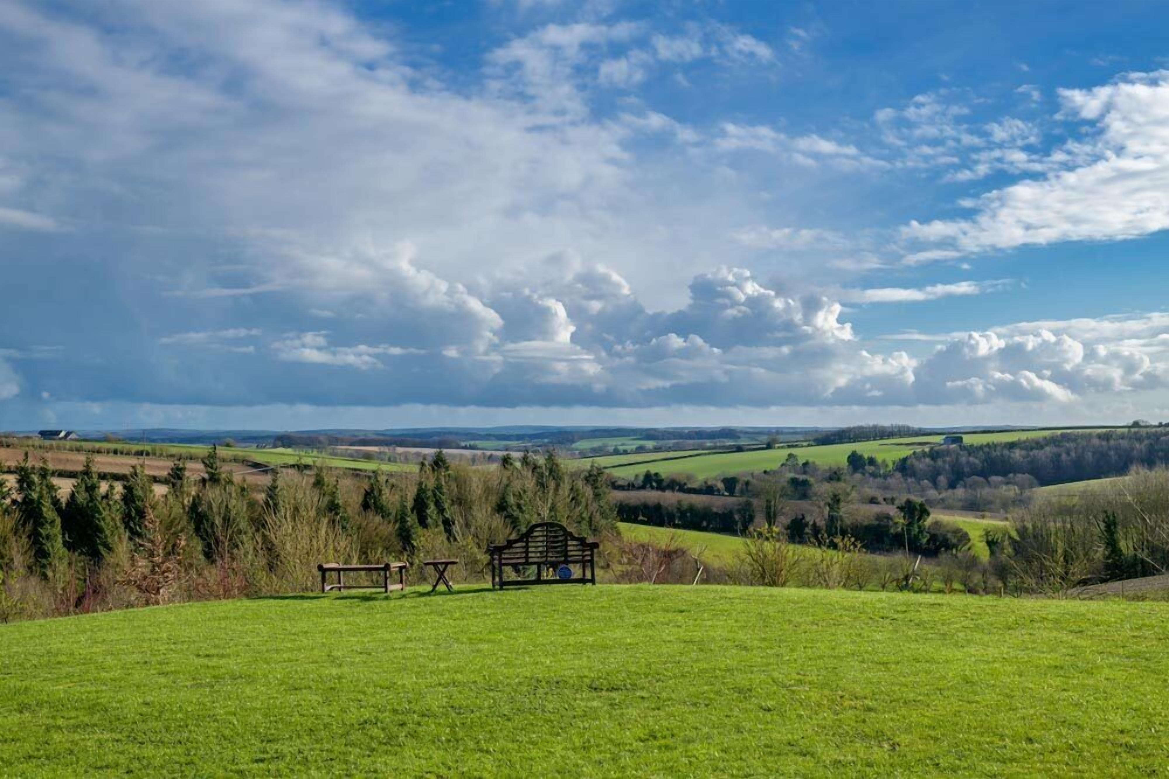 Um campo gramado com um banco e uma mesa, com vista para uma paisagem rural com campos e árvores.