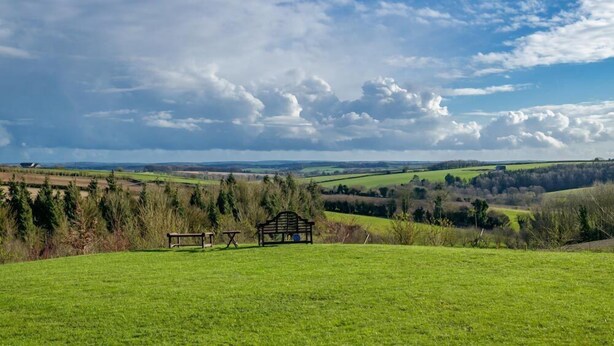 Um campo gramado com um banco e uma mesa, com vista para uma paisagem rural com campos e árvores.