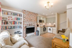 Ein Wohnzimmer mit Kamin, Bücherregal, Sofa, Couchtisch und Küchenzeile mit Geräten.