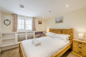 A bedroom with a wooden bed, a bedside table with a lamp, a window with curtains, a bookshelf, and a painting on the wall. - 3 Bed in Whitby (oc-c40269) (Whitby)