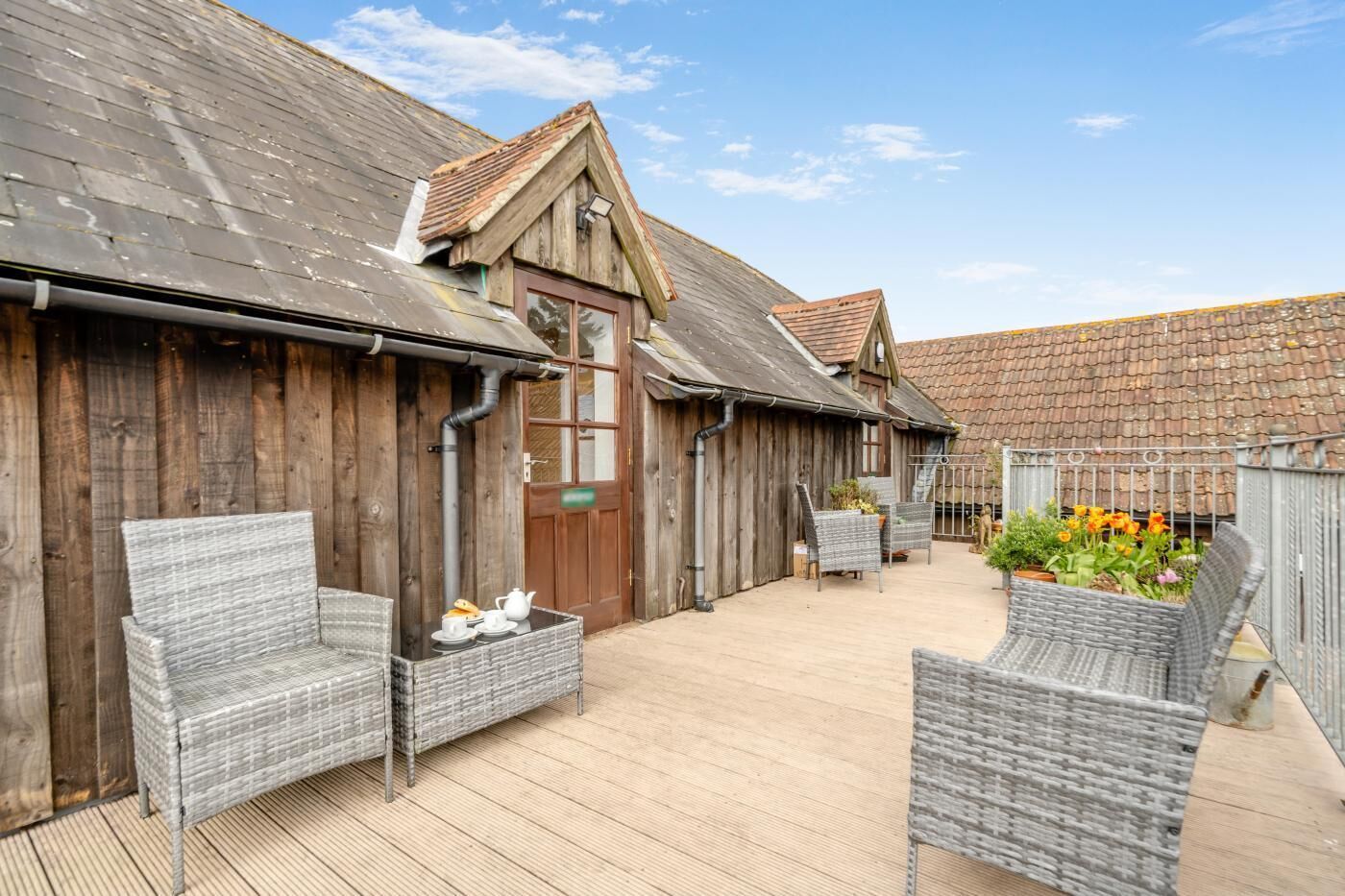 A wooden building with a tiled roof, a door, and a small garden area with wicker furniture and potted plants.