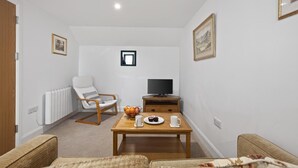 Ein Wohnzimmer mit einem weißen Heizkörper, einem Holz-TV-Schrank, einem Fernseher, einem Holztisch mit Tassen und einer Obstschale, einem weißen Sessel und einem Sofa.