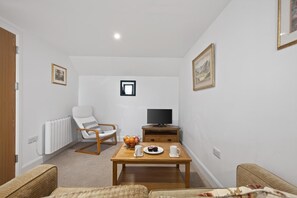 A living room with a white radiator, a wooden TV stand, a television, a wooden coffee table with cups and a fruit bowl, a white armchair, and a sofa. - 1 Bed in Milton Abbas (oc-o35015) (Milton Abbas)
