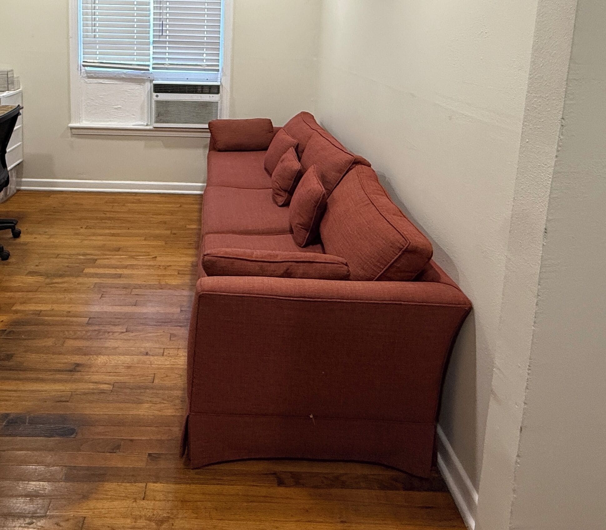 Una esquina de una habitación con piso de madera, un sofá rojo y una ventana con persianas.