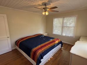 Un dormitorio con una cama, un ventilador de techo, una ventana con persianas y una puerta.