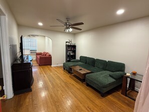 Uma sala de estar com sofá em forma de L, mesa de centro, estante de livros e ventilador de teto.