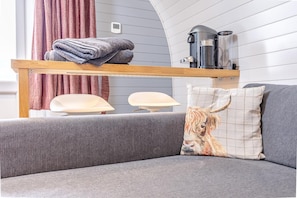 Ein modernes Wohnzimmer mit einem grauen Sofa, einem Holzregal mit gefalteten Handtüchern, einer Kaffeemaschine und einem an der Wand montierten Thermostat.