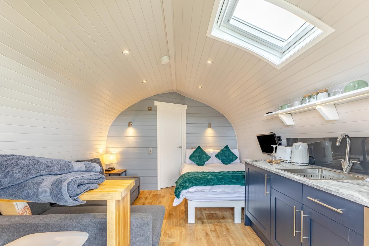 A compact room with a bed, a small table, a bench, a kitchenette, and a skylight.