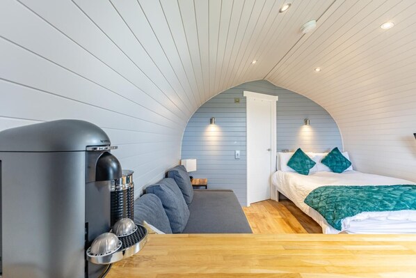 A compact, modern bedroom with a curved ceiling, a bed with teal bedding, a seating area with a sofa, and a kitchenette with a coffee machine.
