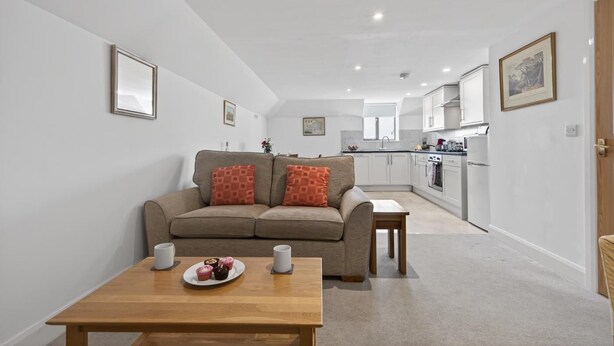 A modern living room with a sofa, a dining table with a plate of food, and a kitchen in the background. - 1 Bed in Milton Abbas (oc-v35014) (Milton Abbas)