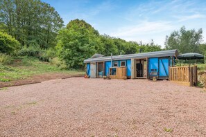 A rustic cabin with a gravel driveway, wooden deck, and a covered outdoor seating area. - 1 Bed in Longhope (oc-m45889) (Longhope)