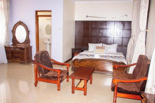 A bedroom with a large bed, two wooden armchairs, a small wooden table, a nightstand, a mirror, and a bathroom with a sink and mirror.