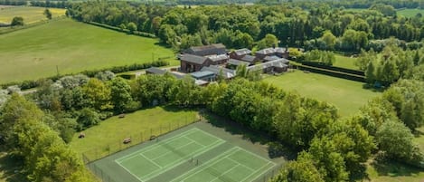 A residential property with a tennis court, surrounded by lush greenery and a large house with multiple buildings.