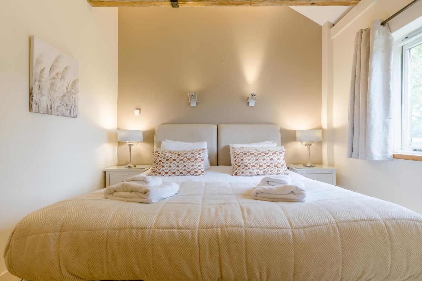 A double bed with a beige quilt, two bedside tables with lamps, patterned pillows, and a window with curtains.
