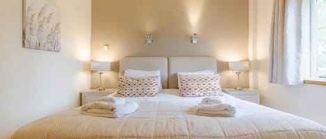 Una cama doble con colcha color beige, dos mesitas de noche con lámparas, almohadas estampadas y una ventana con cortinas.