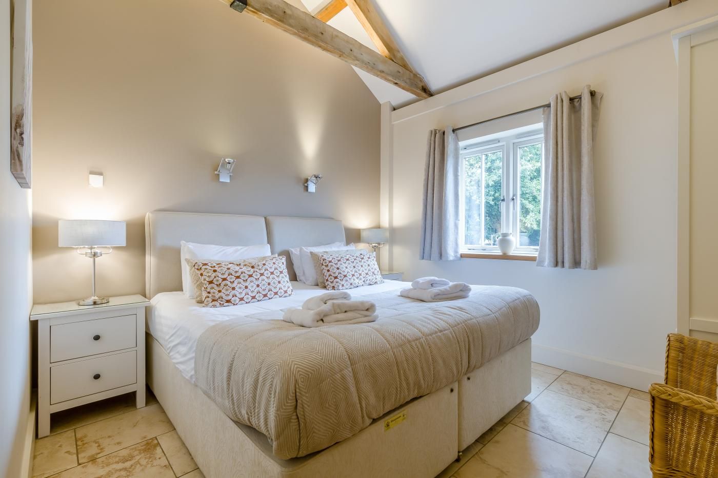 Quarto com cama, mesas de cabeceira, janela com cortinas e um vigamento de madeira no teto.