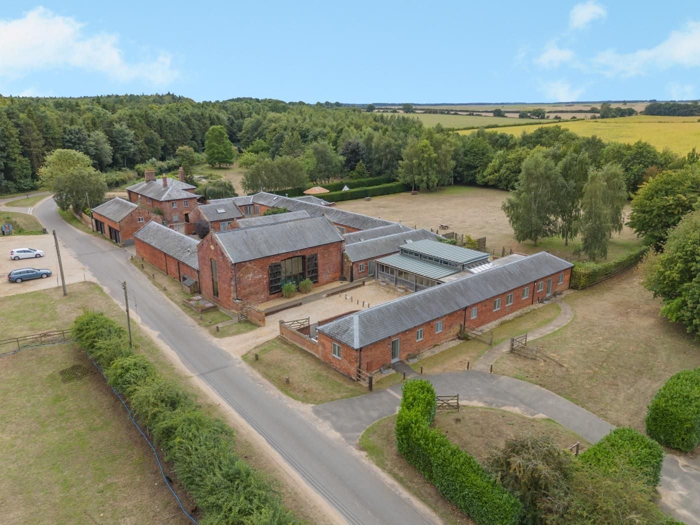 A large brick building with multiple sections, surrounded by a green landscape and a road.