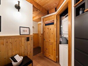 A wooden interior with a door, a white wall with a framed sign, a black table with a basket, and a white appliance. - Escape to Ptarmigan Village 36 (Whitefish)