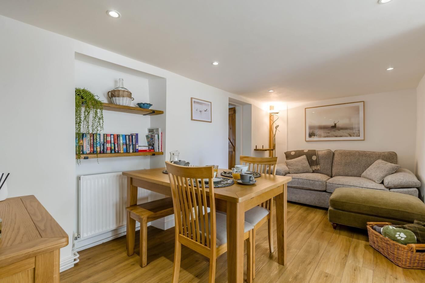 A cozy living room with a wooden dining table, chairs, a sofa, and a bookshelf with books and decorative items.