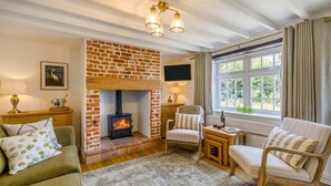 A cozy living room with a brick fireplace, comfortable seating, a television, and a large window with curtains. - 2 Bed in Great Massingham (oc-a45882) (Great Massingham)
