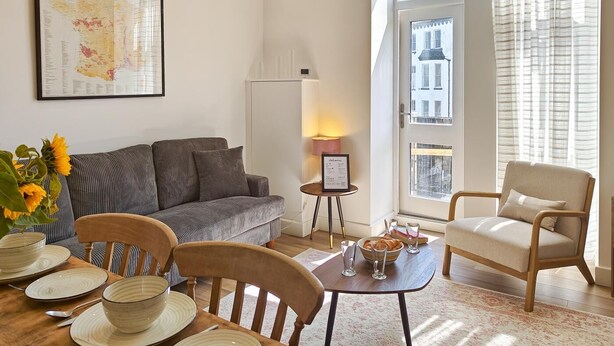 A living room with a sofa, chairs, a table with plates and glasses, a lamp, and a window with a view of buildings. - 2 Bed in Ilfracombe (oc-b45841) (Ilfracombe)