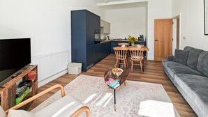 A modern living room with a flat-screen TV, a grey sofa, wooden chairs, a coffee table, and a kitchen area with a dining table. - 2 Bed in Ilfracombe (oc-b45841) (Ilfracombe)