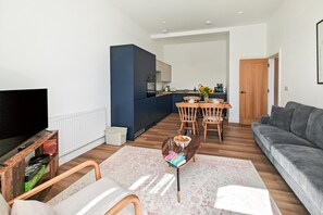 Ein modernes Wohnzimmer mit Flachbildfernseher, einem grauen Sofa, Holzstühlen, einem Couchtisch und einer Küchenzeile mit Esstisch.