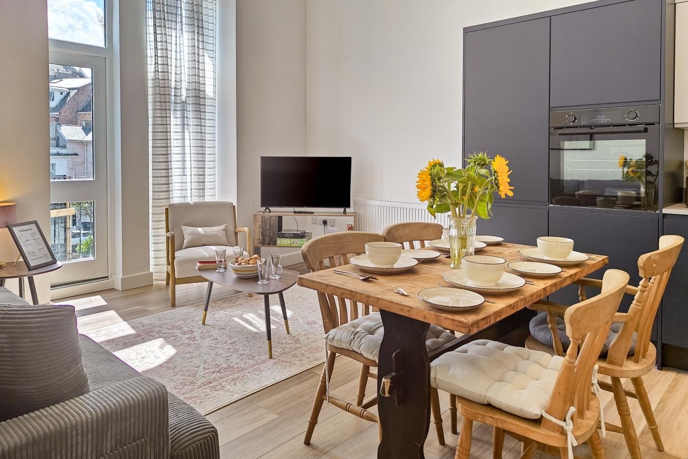 A modern living room with a dining area, a flat-screen TV, and a kitchen with a built-in oven.
