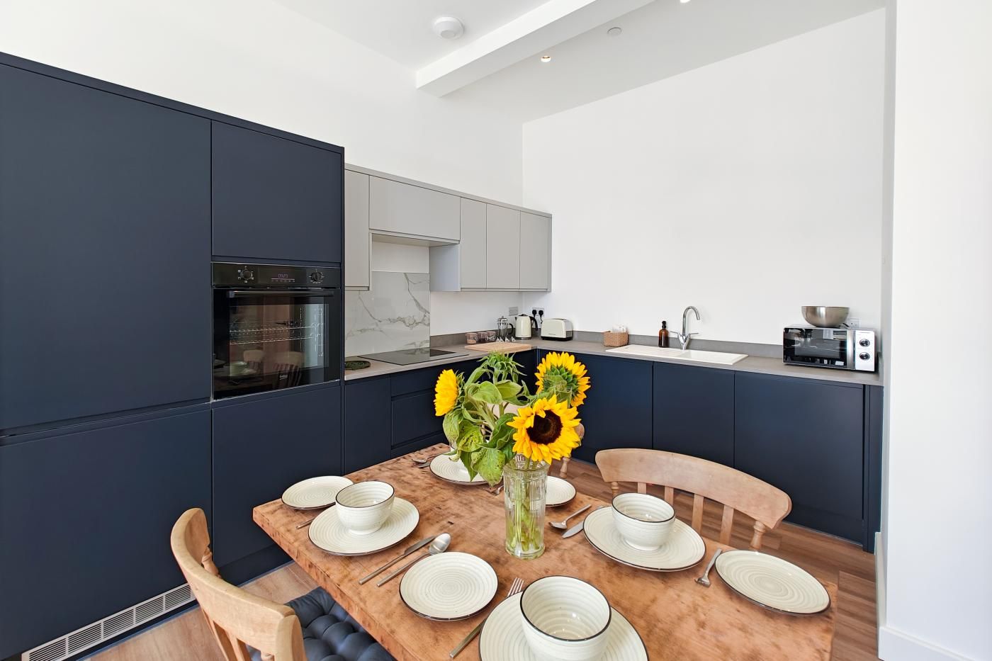 A modern kitchen with dark cabinetry, a built-in oven, and a dining area with a wooden table set for four.
