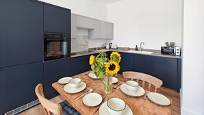 A modern kitchen with dark cabinetry, a built-in oven, and a dining area with a wooden table set for four. - 2 Bed in Ilfracombe (oc-b45841) (Ilfracombe)
