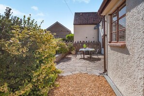 Eine Terrasse mit Tisch und Stühlen, ein gepflasterter Weg und ein Heckenstreifen.