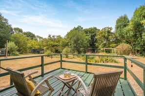 Une terrasse en bois, agrémentée d’une table et de chaises, entourée d’une rambarde verte, et offrant une vue sur un jardin avec des arbres et un étang.