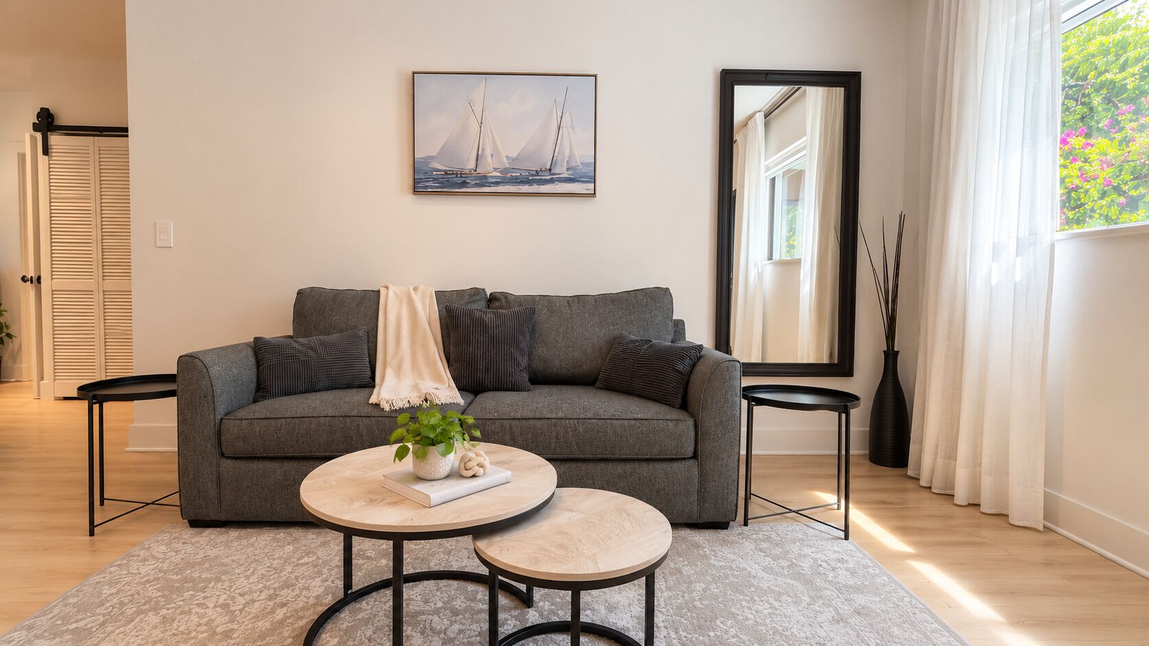En moderne stue med en grå sofa, to runde sofaborde, en plante og et maleri af en sejlbåd på væggen.
