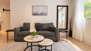 Een moderne woonkamer met een grijze bank, twee ronde salontafels, een plant en een schilderij van een zeilboot aan de muur.