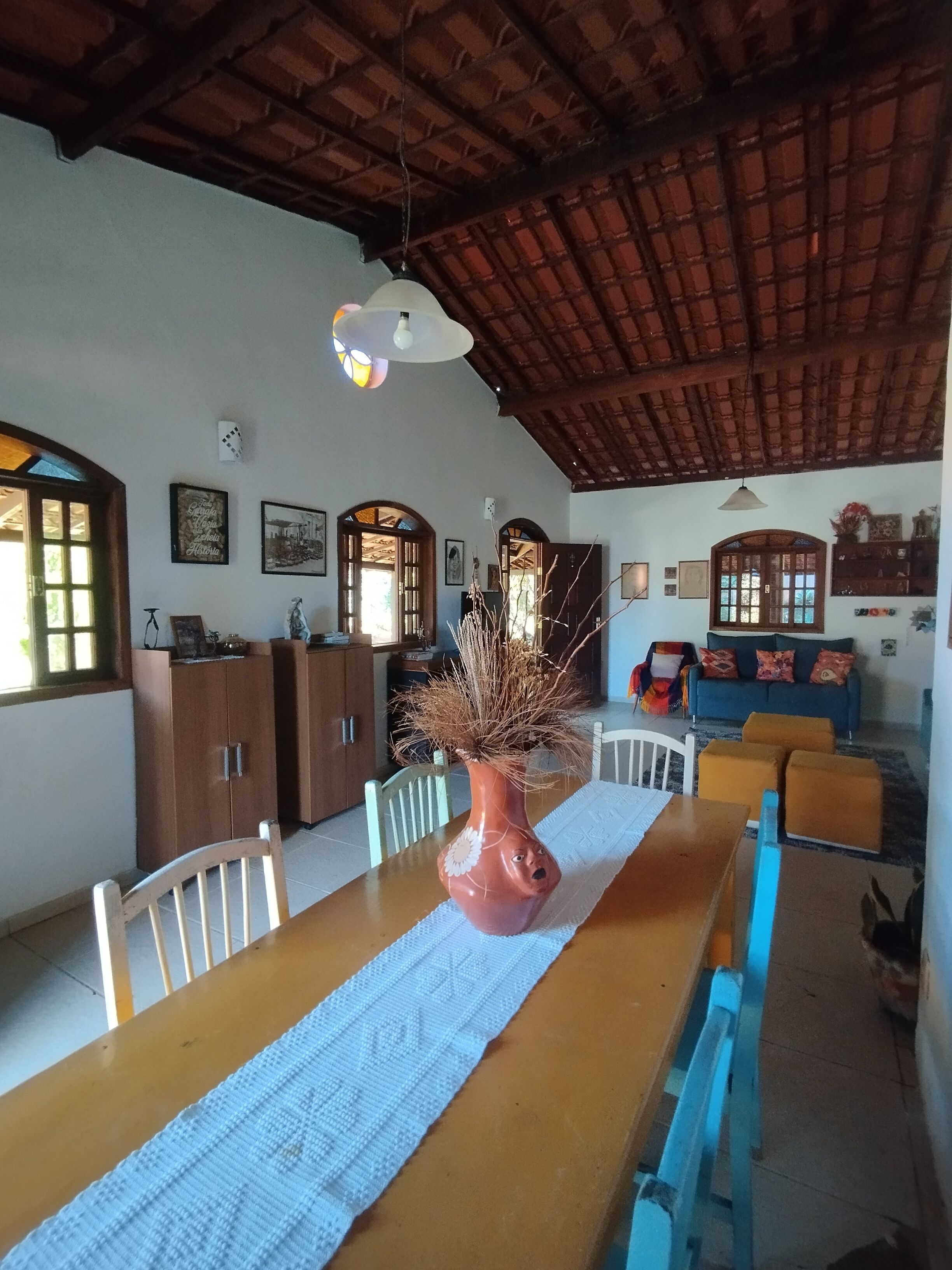 Un comedor con una larga mesa de madera, sillas, un jarrón con ramas secas y un techo con vigas a la vista.