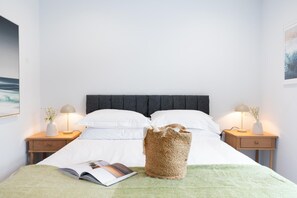 A neatly made bed with white linens, two bedside lamps, a woven basket, and a book on the bed. - 2 Bed in Falmouth (oc-b669263) (Flushing)