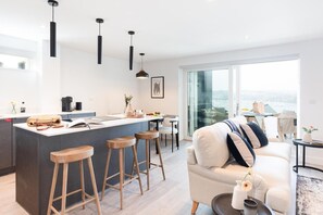 Eine moderne Küche mit einer zentralen Kochinsel, Barhocker, einem Essbereich und einem Wohnbereich mit Sofa und Blick ins Freie.
