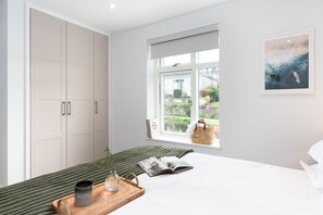 Ein Schlafzimmer mit großem Kleiderschrank, einem Fenster mit Ausblick, einem Nachttisch mit Zeitschrift und einem gerahmten Kunstwerk an der Wand.