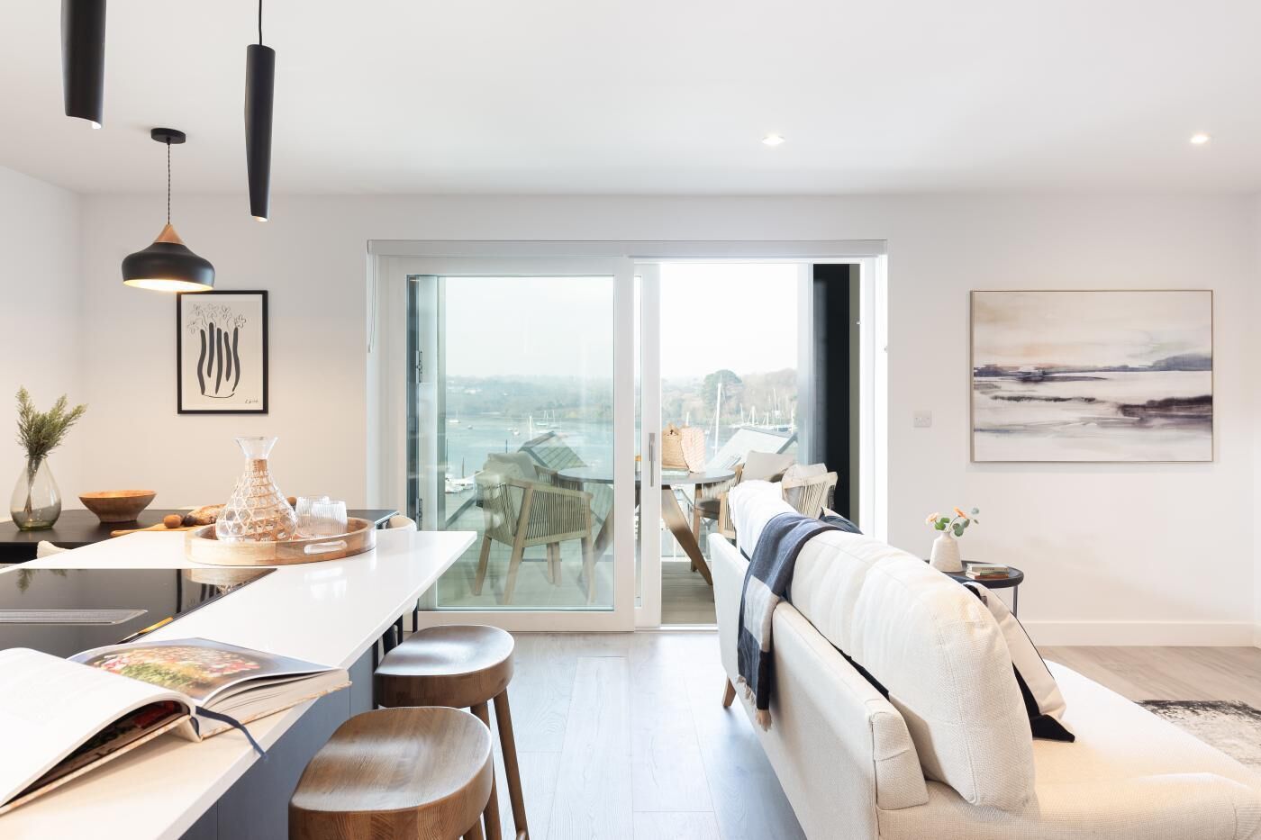 A modern living room with a sofa, dining table, and chairs, a large window with a view of the sea, and a painting on the wall.