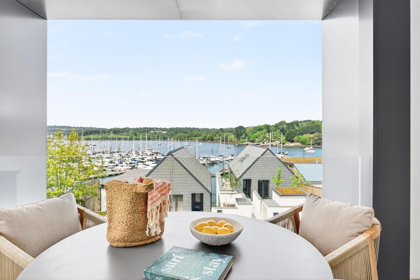 A balcony with a view of a marina, wicker basket, bowl of fruit, and a book.