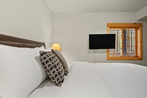 A hotel room with a bed, a TV mounted on the wall, a window with blinds, and a bedside lamp.