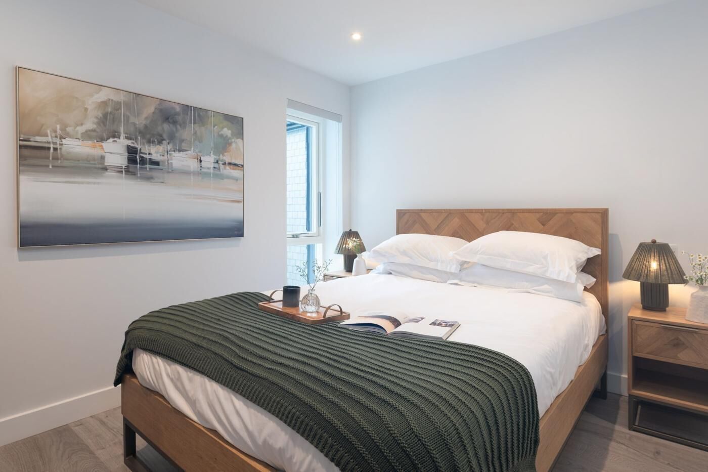 Ein ordentlich bezogenes Bett mit Holz Kopfteil, ein Nachttisch mit Lampe und ein großes abstraktes Gemälde an der Wand.