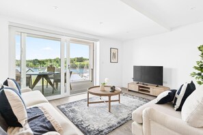 A modern living room with a large TV, a sofa, a coffee table, and a view of a deck with outdoor furniture. - 2 Bed in Falmouth (oc-b669265) (Flushing)