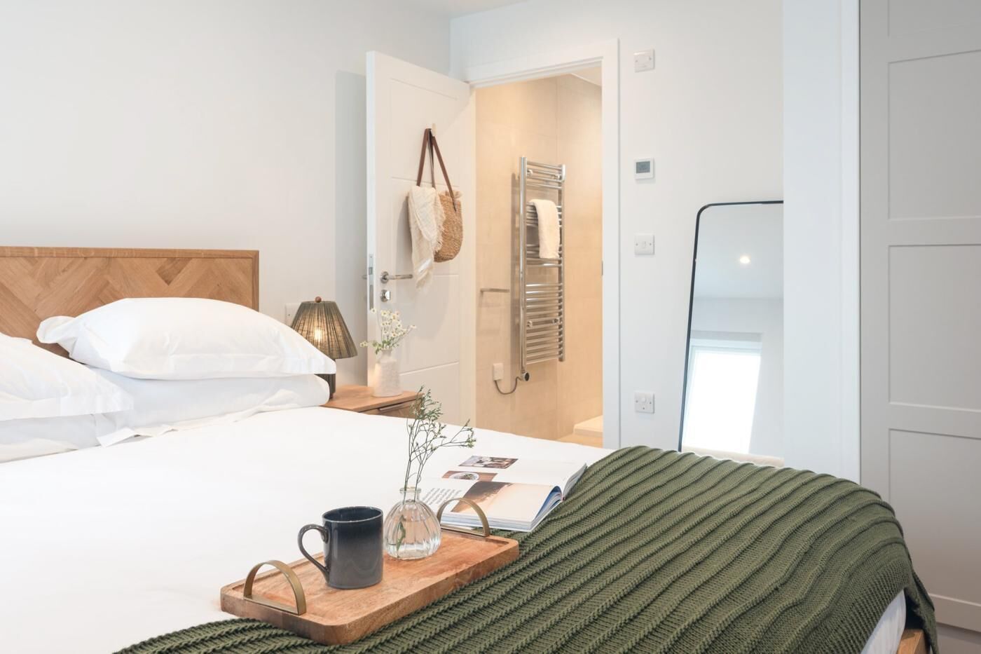 A hotel room with a bed, a bedside table with a lamp, a tray with a mug and a vase, and a door leading to a bathroom.