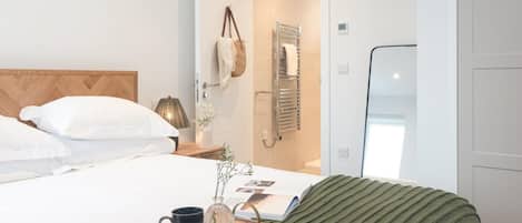 A hotel room with a bed, a bedside table with a lamp, a tray with a mug and a vase, and a door leading to a bathroom.