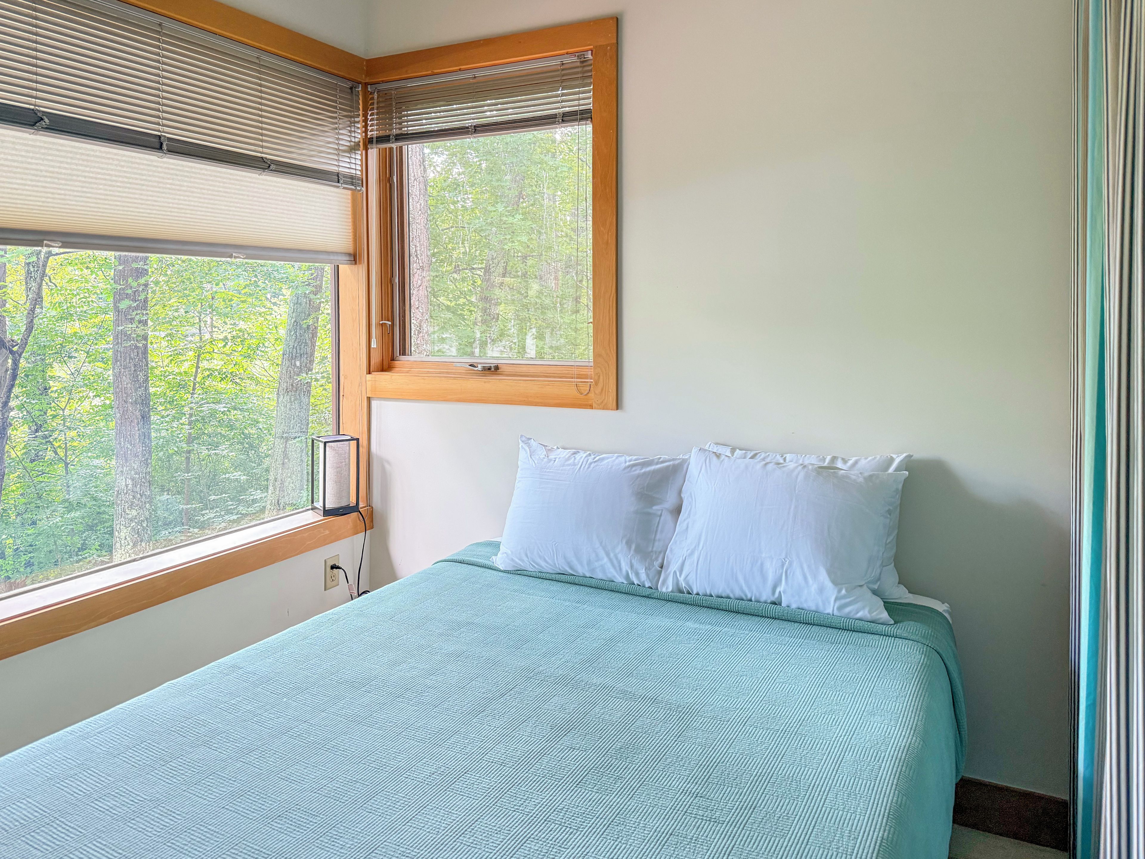 Ein Schlafzimmer mit einem Bett, einem Fenster mit Jalousien und Blick auf Bäume.